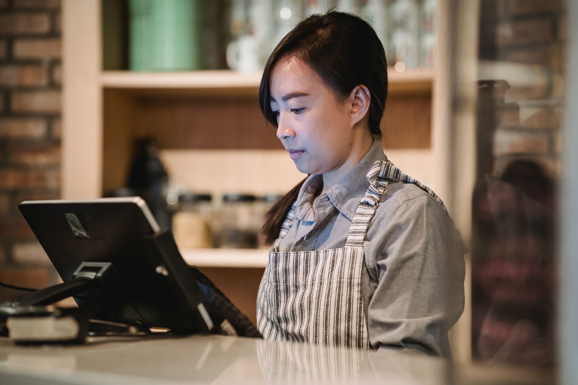 A barista in a striped apron working diligently at a cafe counter, using a point-of-sale system.